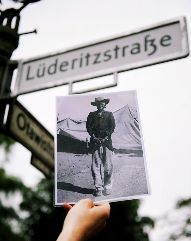 Ein Aktivist hält ein Motiv aus der deutschen Kolonialzeit hoch. Im Hintergrund ist das Straßenschild der Lüderitzstraße im Berliner Wedding zu sehen, die in Cornelius-Fredericks-Straße umbenannt werden soll.
