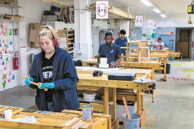 Alina Eilenstein ölt Leisten für eine Holzhandtasche.  Mit ihr im Werkstattraum arbeiten Bootsbau-Azubi Kalipha Keita mit der CNC-Fräse und Mirwais Ahmadzai an einem Hängeschrank.
