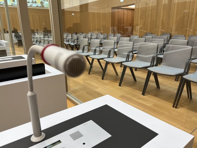 Ein Mikrofon ist an einem Tisch in dem Gerichtssaal im Oberlandesgericht angebracht, in dem der Reichsbürger-Prozess verhandelt wird. Die Angeklagten sitzen hinter Trennwänden aus Glas.
